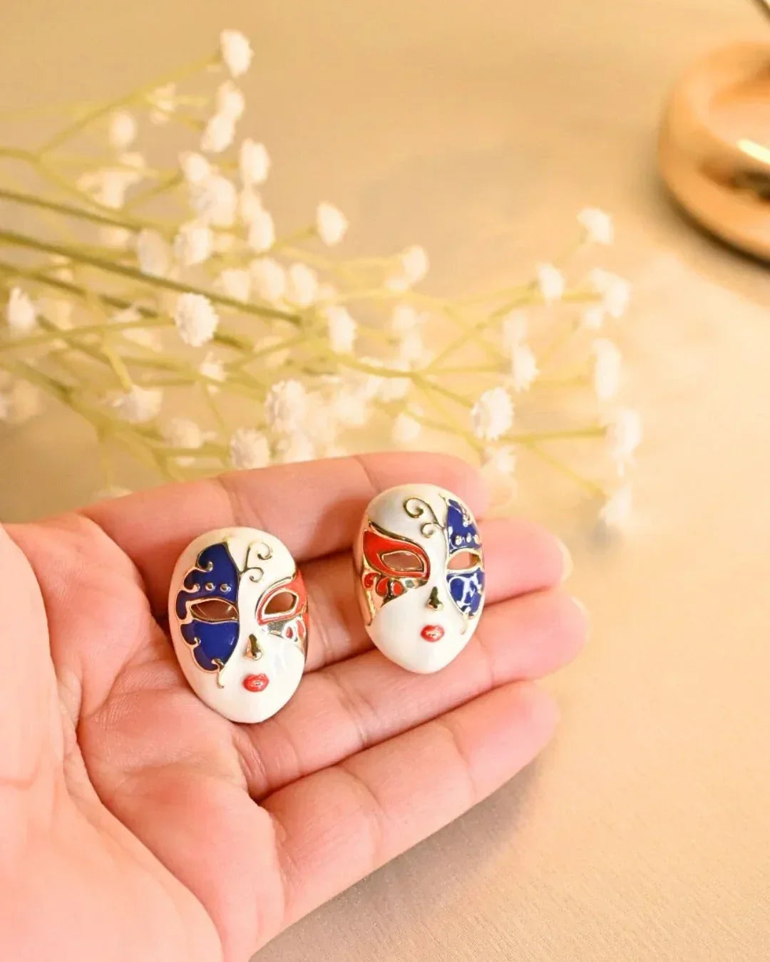 Hand holding two colorful Venetian mask earrings with red and blue enamel design