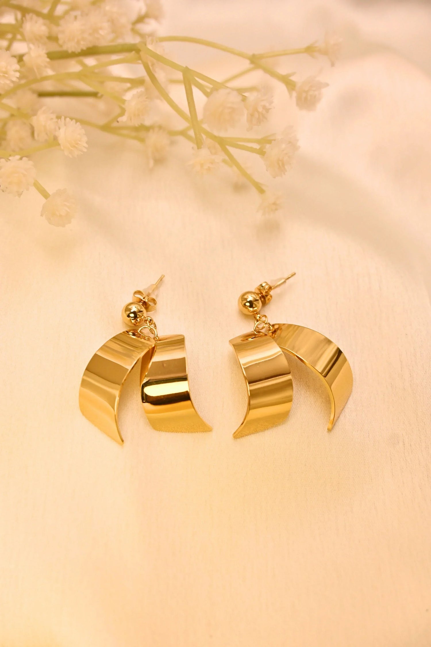 Gold-tone statement earrings with curved design on soft beige fabric, floral accent background