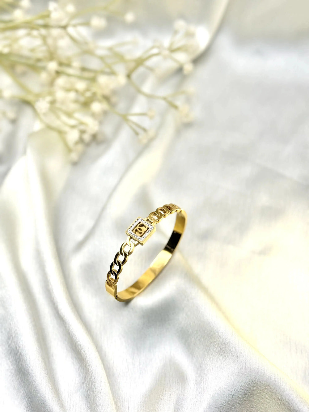 Gold chain bracelet with square charm on white satin fabric, decorative flowers in background