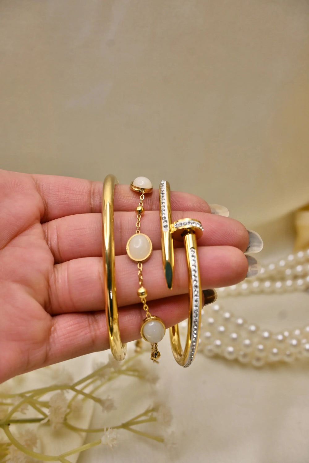 Hand holding gold bangle, pearl bracelet, and diamond-studded nail-shaped bracelet jewelry