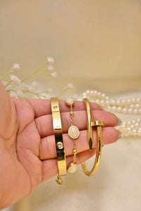 Hand holding three gold bracelets with gemstones, pearls and floral background