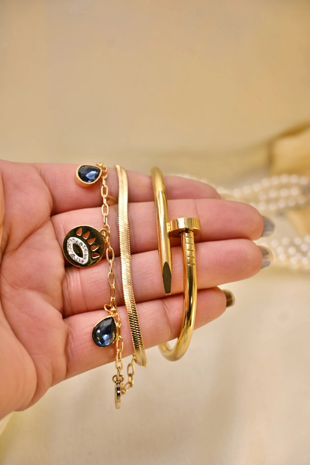Hand holding gold bracelets and a gold chain with blue gemstone charms and a modern nail-shaped bangle