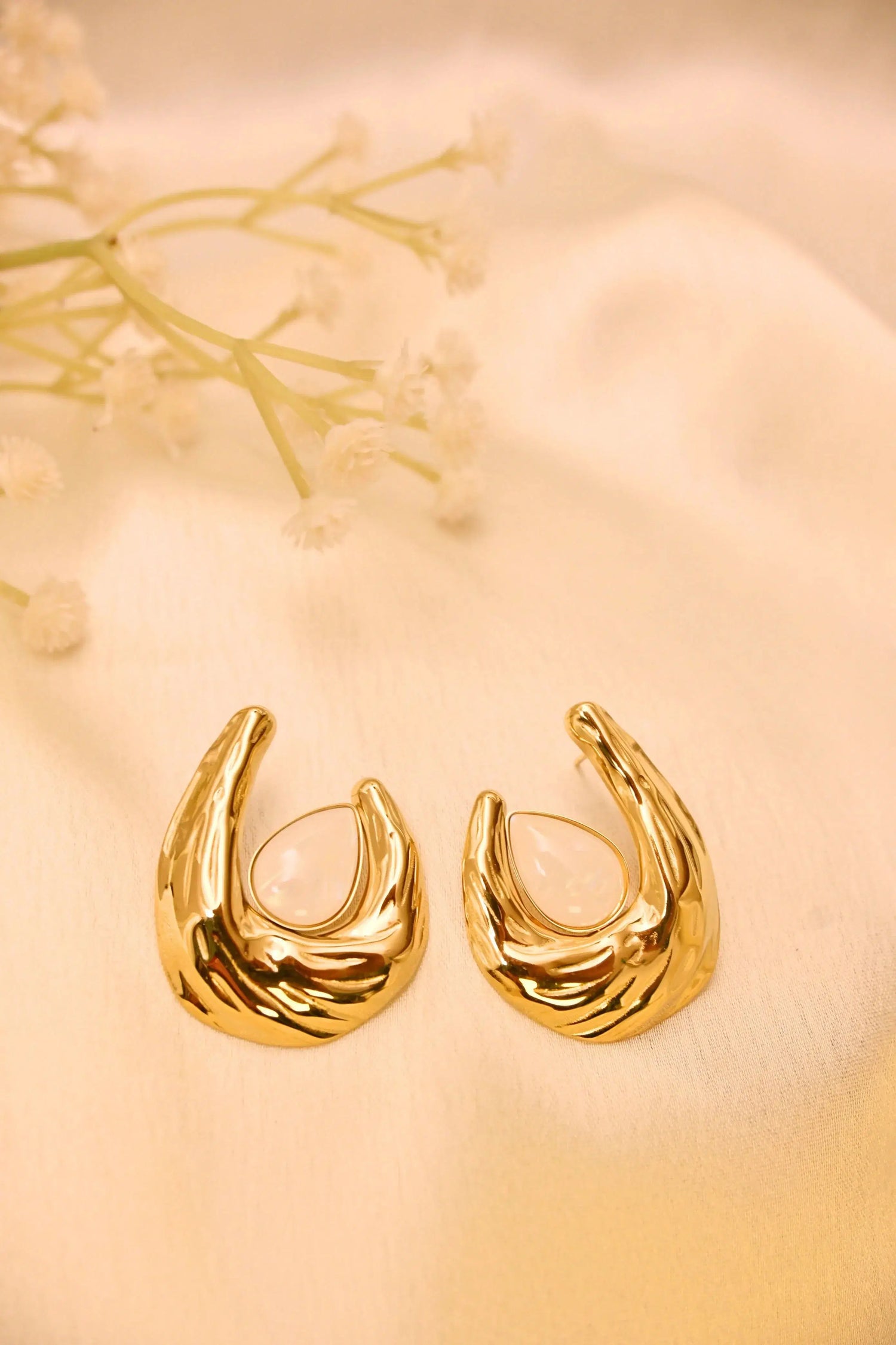 Gold-plated earrings with teardrop white stones on a soft beige fabric background, floral accent