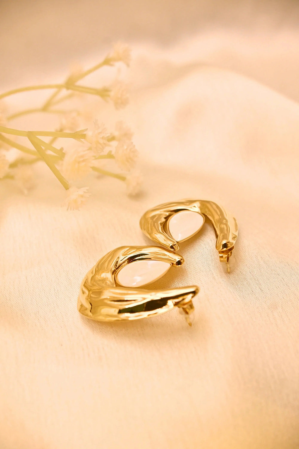 Gold-tone abstract stud earrings with white inlay on a soft beige fabric, floral accent nearby