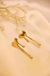 Gold heart-shaped drop earrings on soft beige fabric with delicate white flowers