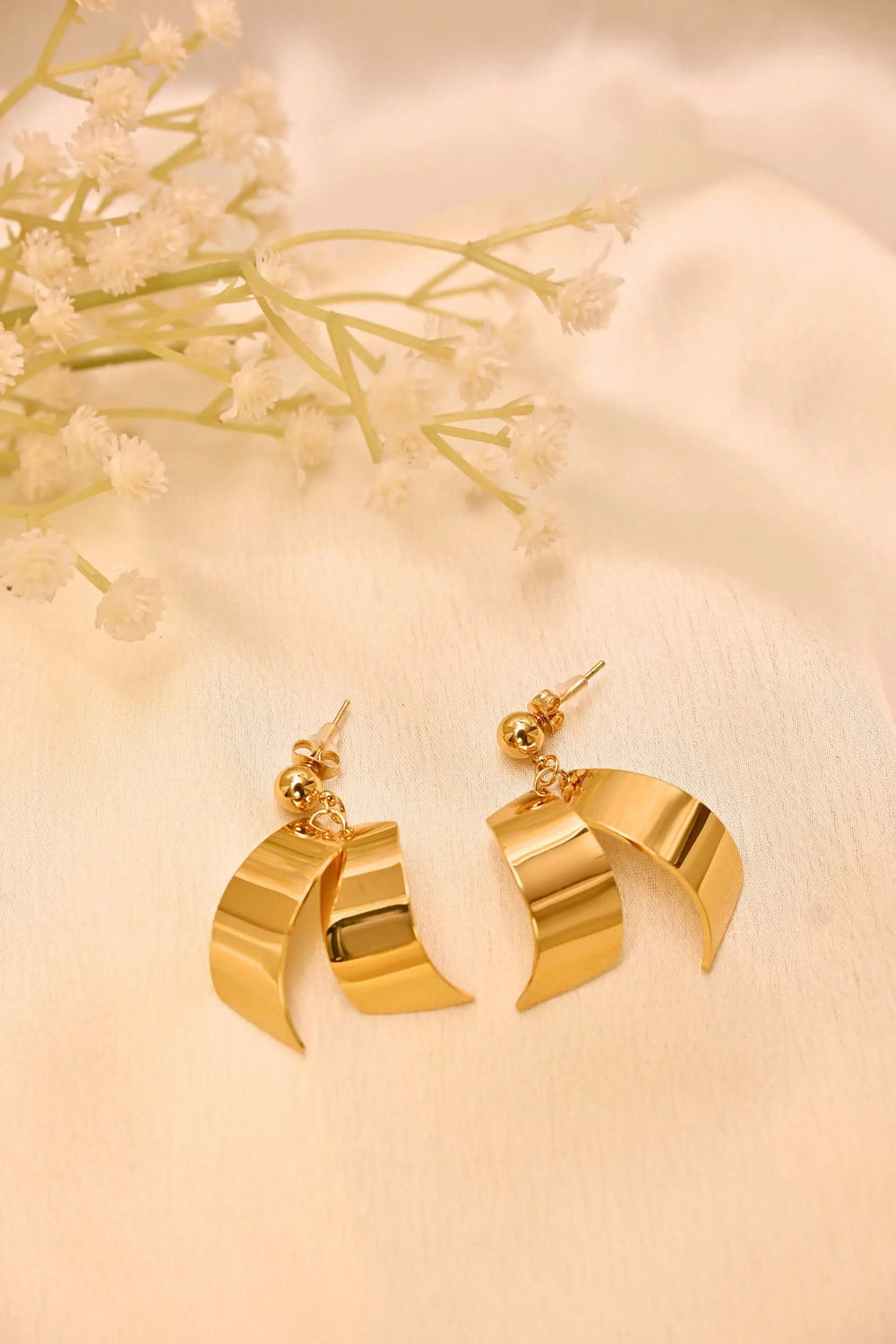 Gold tone curved drop earrings with a modern design on a cream fabric background and flowers