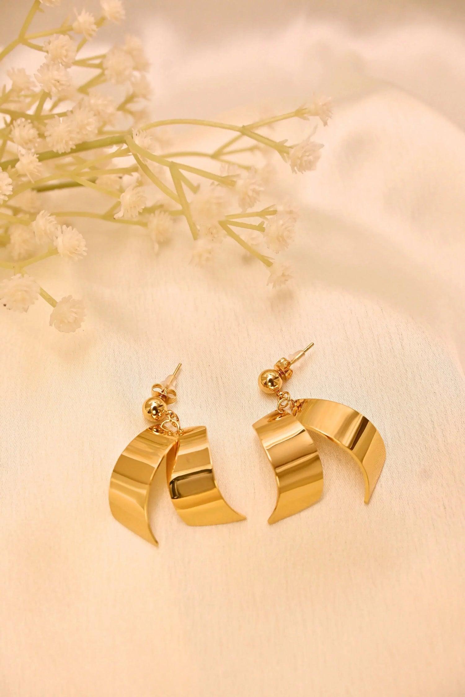 Gold tone curved drop earrings with a modern design on a cream fabric background and flowers