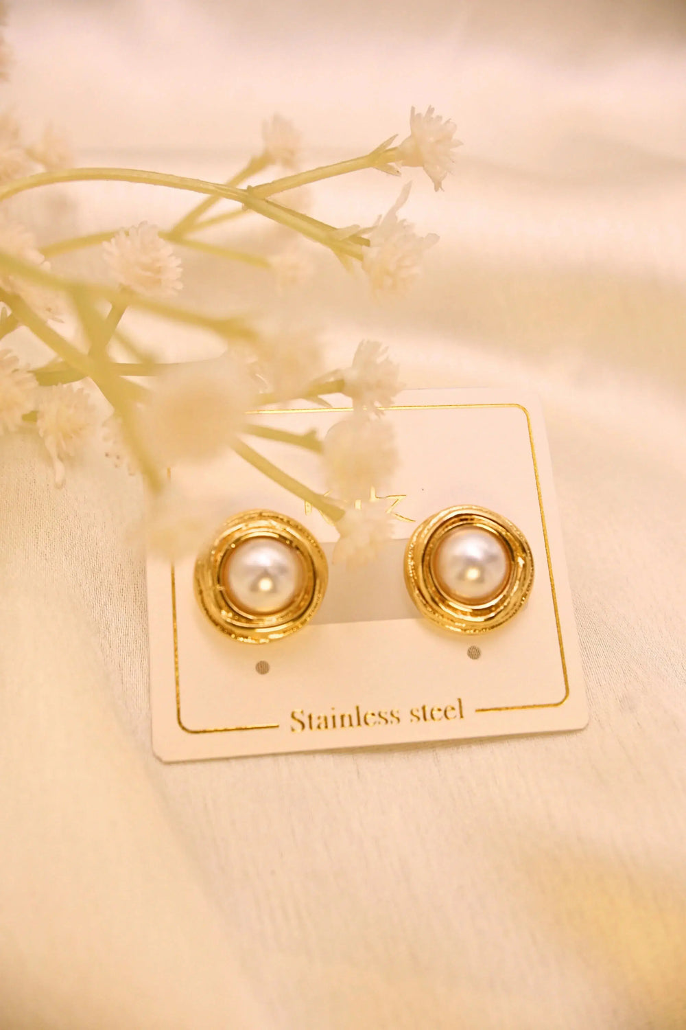 Elegant gold-plated stainless steel pearl stud earrings on a display card with soft floral decor