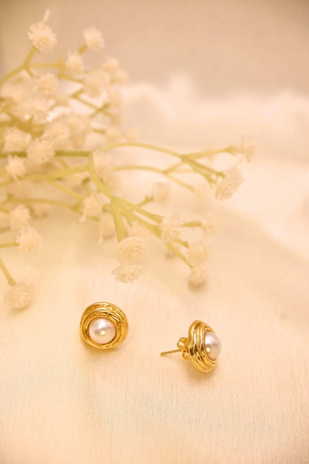 Gold-plated pearl stud earrings on cream fabric with dried white flowers