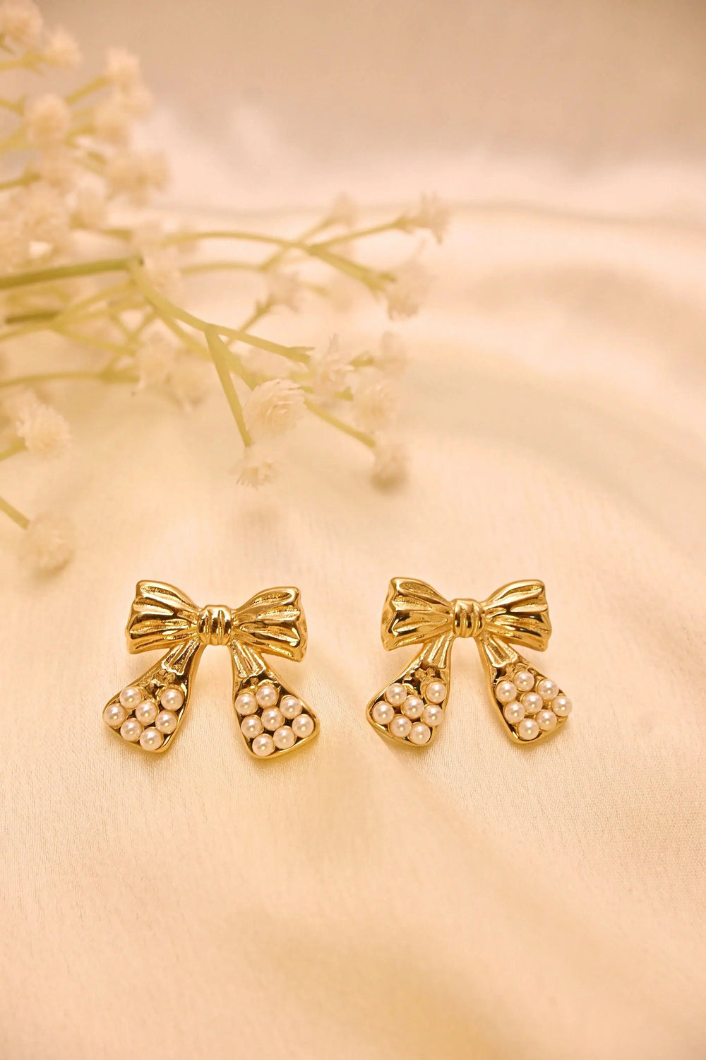 Gold bow earrings with pearl detailing on a cream fabric background