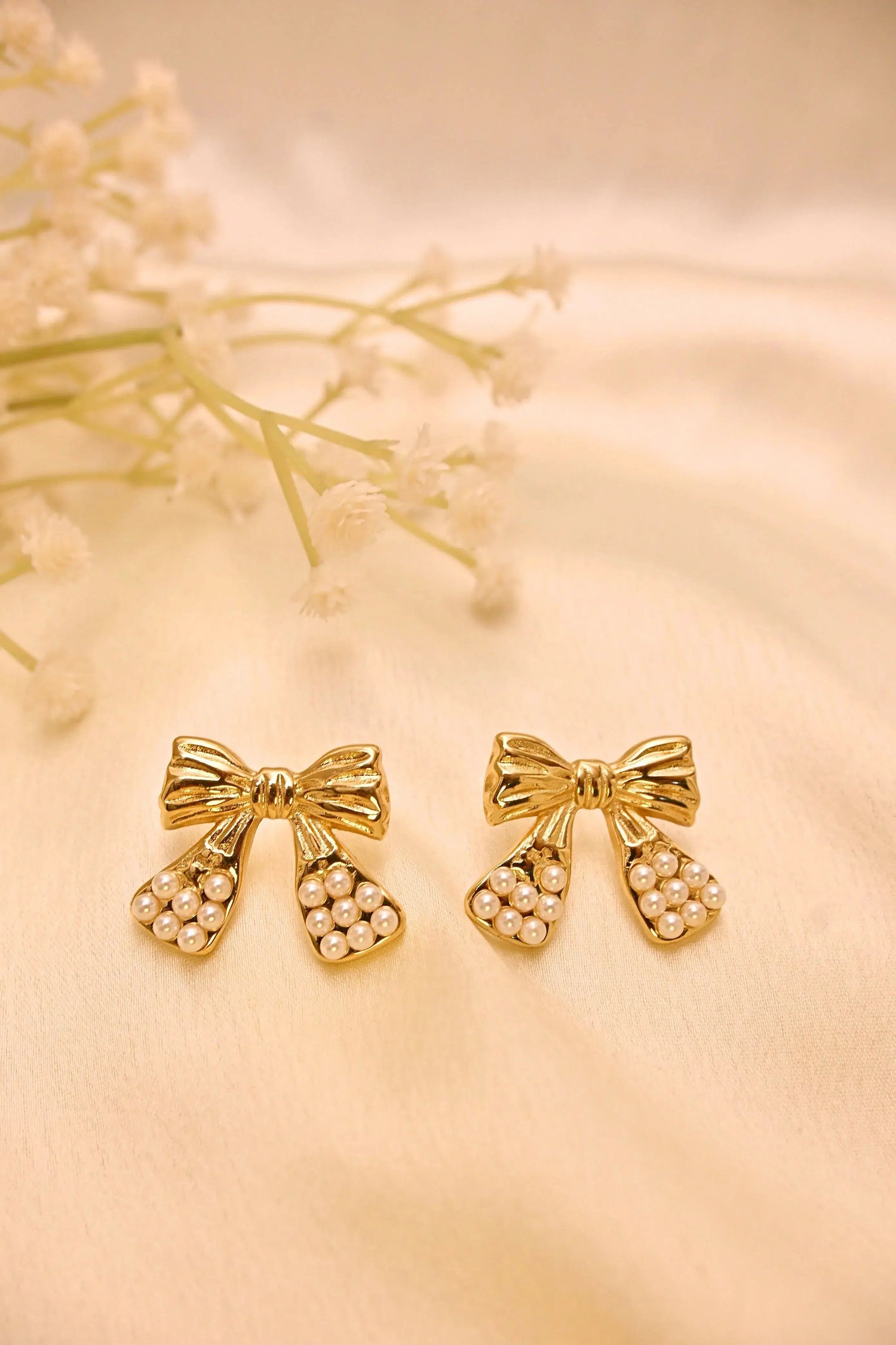Gold bow earrings with pearl detailing on a cream fabric background