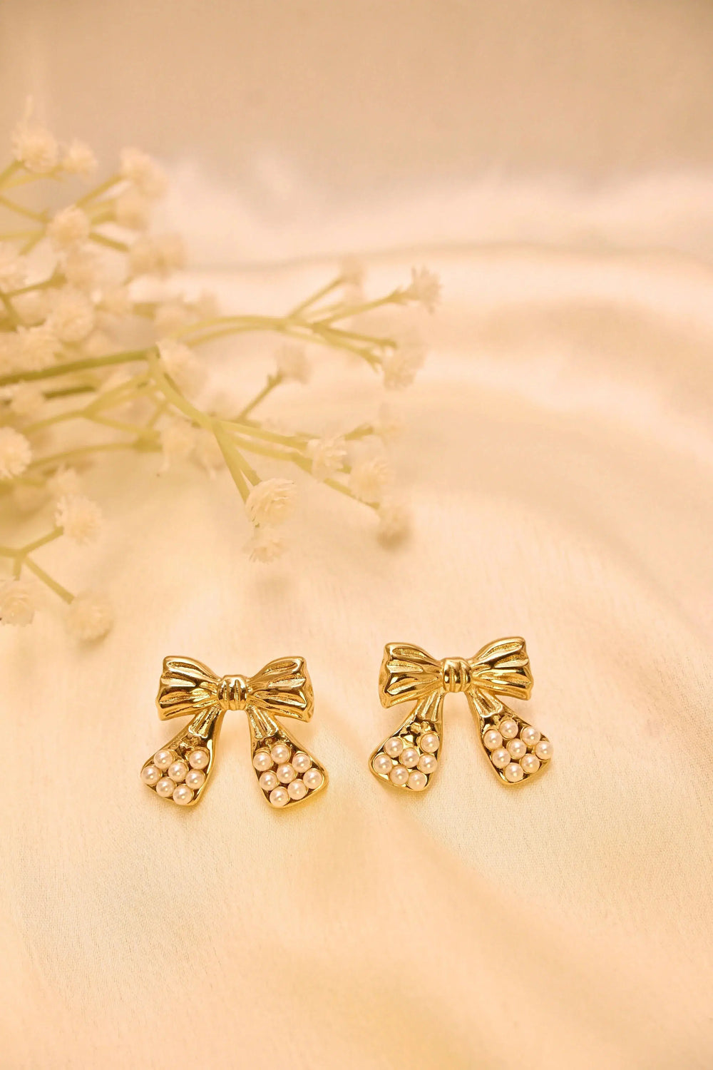Gold bow-shaped stud earrings with pearl accents on a cream satin background