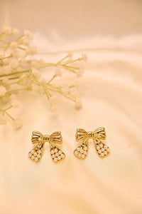 Gold bow-shaped stud earrings with pearl accents on a cream satin background