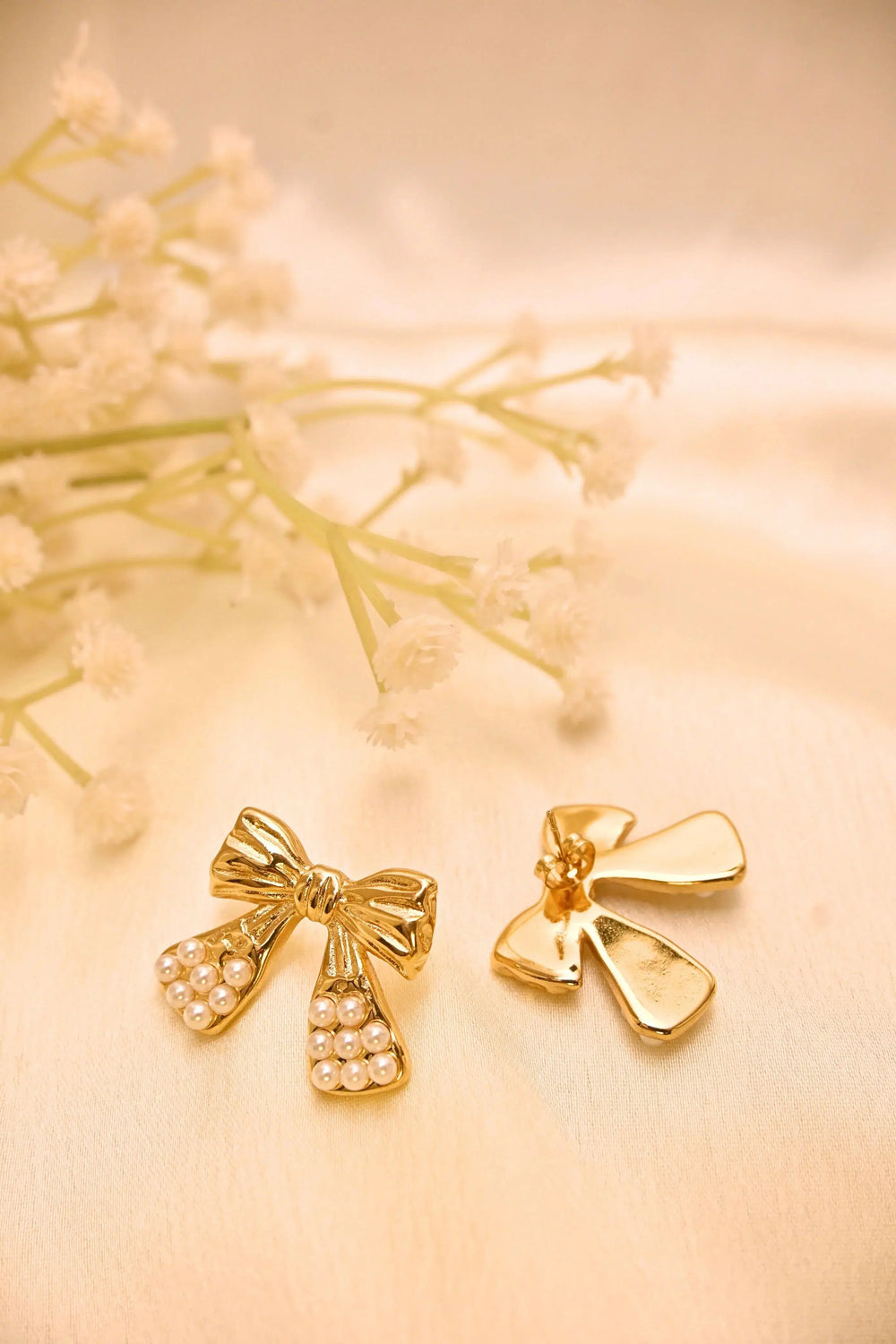 Gold bow earrings with pearl accents on a light fabric background, beside delicate flowers.
