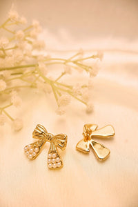 Gold bow earrings with pearl accents on a light fabric background, beside delicate flowers.