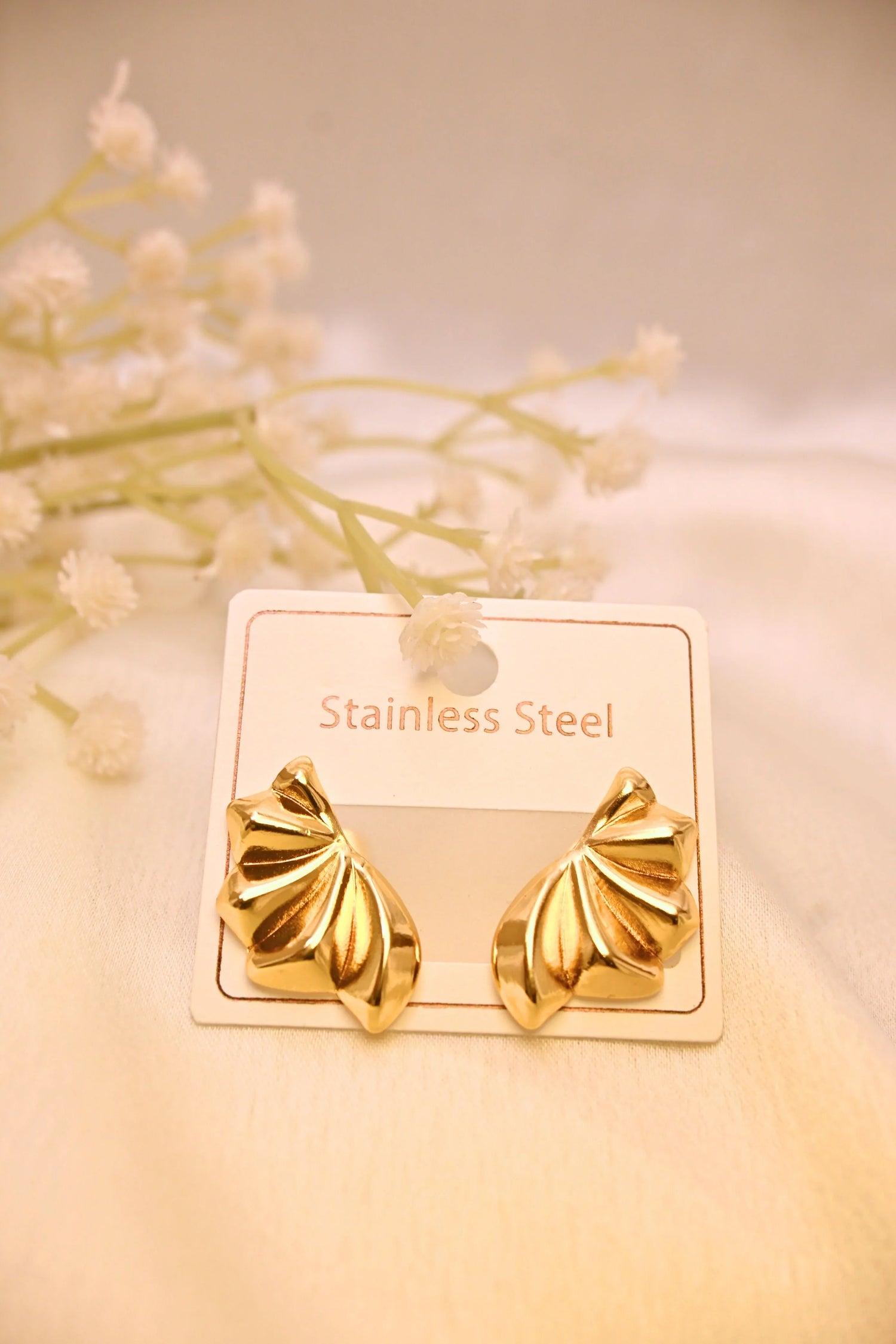 Gold-tone stainless steel leaf-shaped earrings on display card with soft floral background
