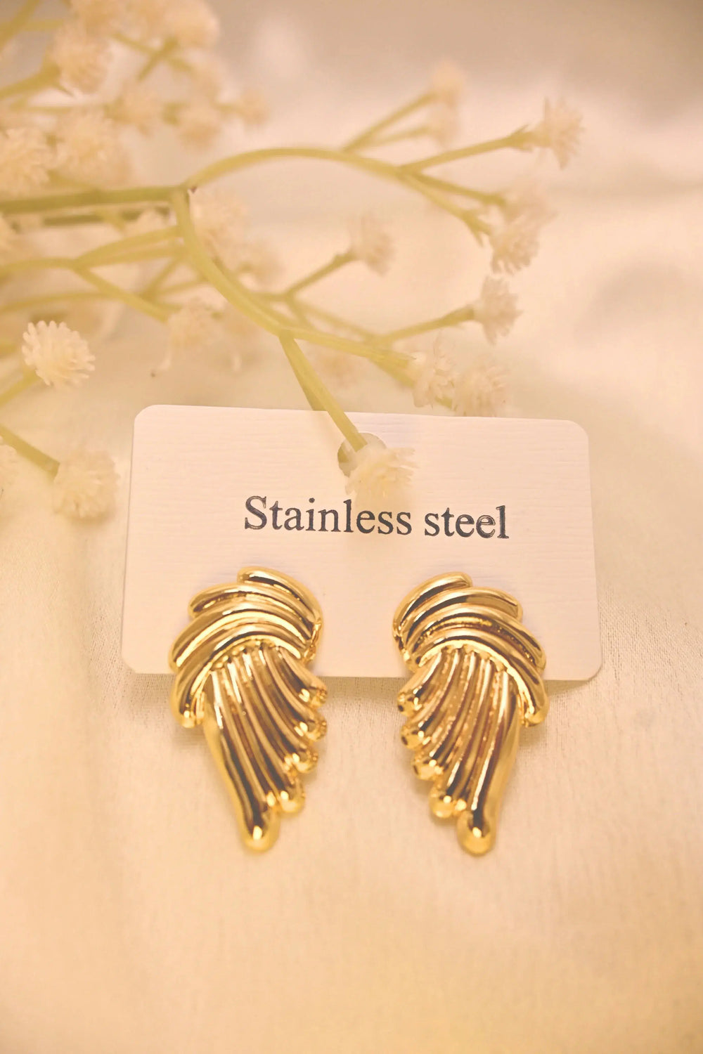 Gold-tone stainless steel wing-shaped earrings on a white display card with flowers.