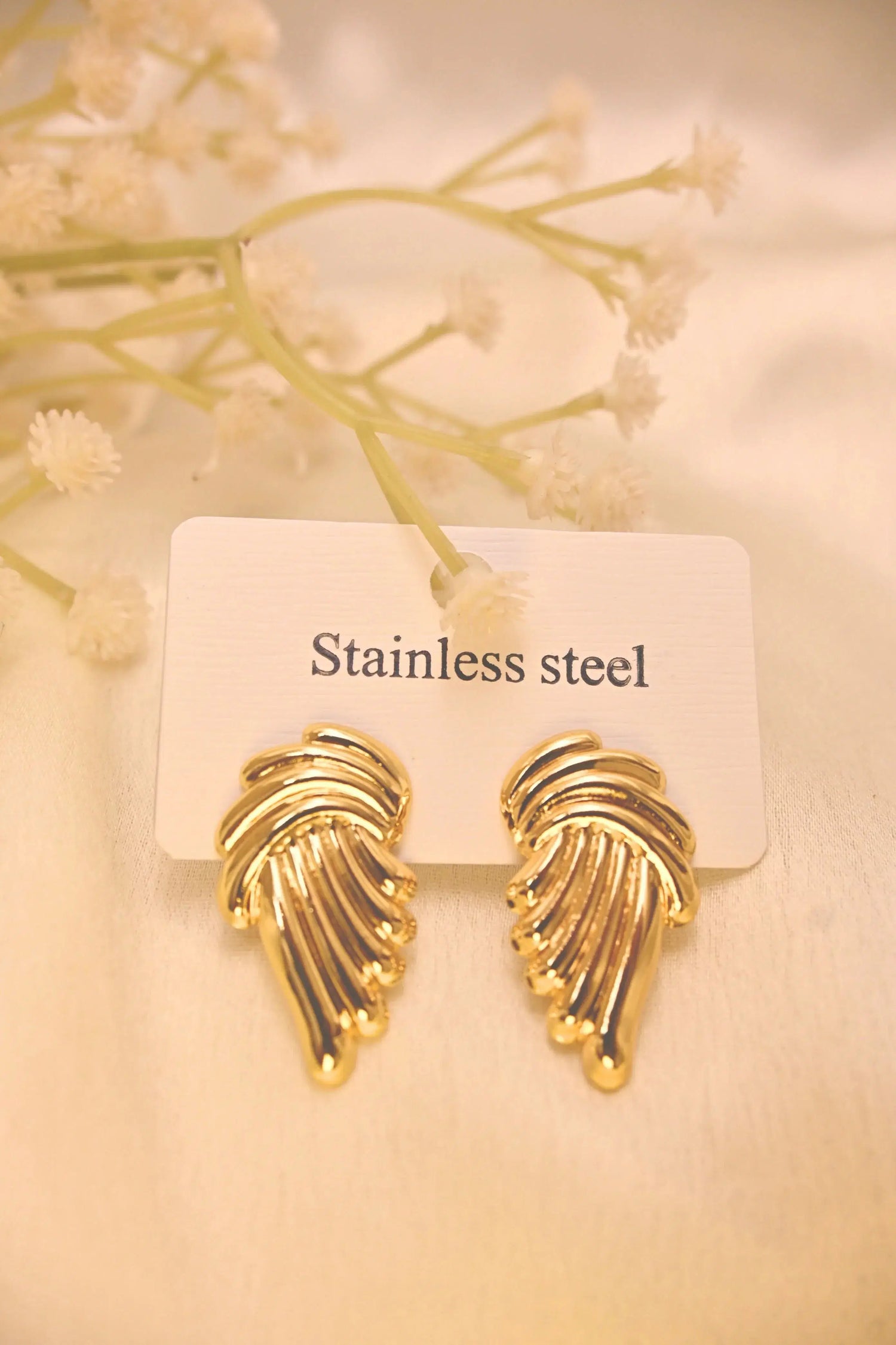 Gold-tone stainless steel wing-shaped earrings on a white display card with flowers.