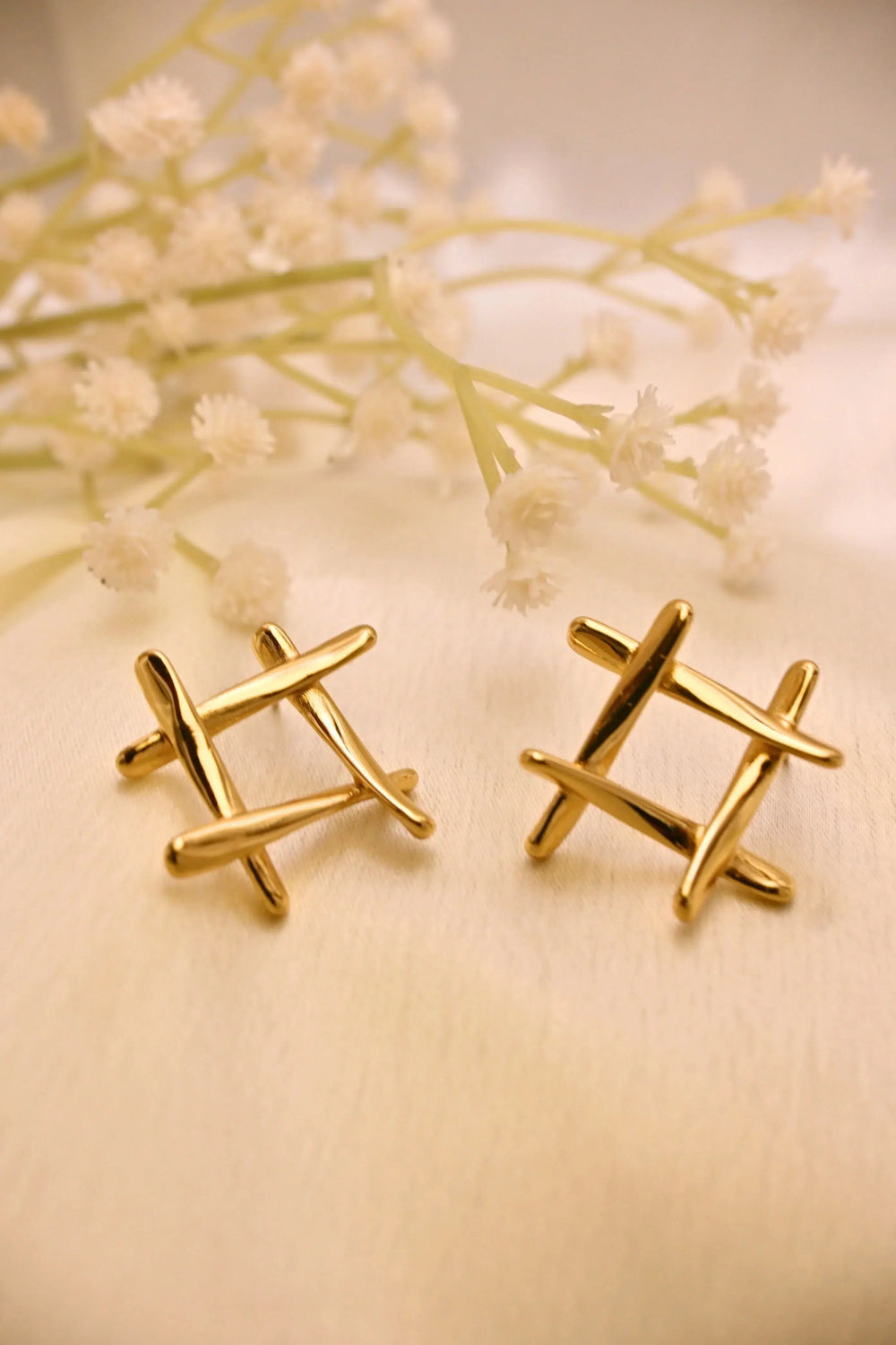Pair of modern gold stud earrings with crisscross design on cream fabric, floral decor