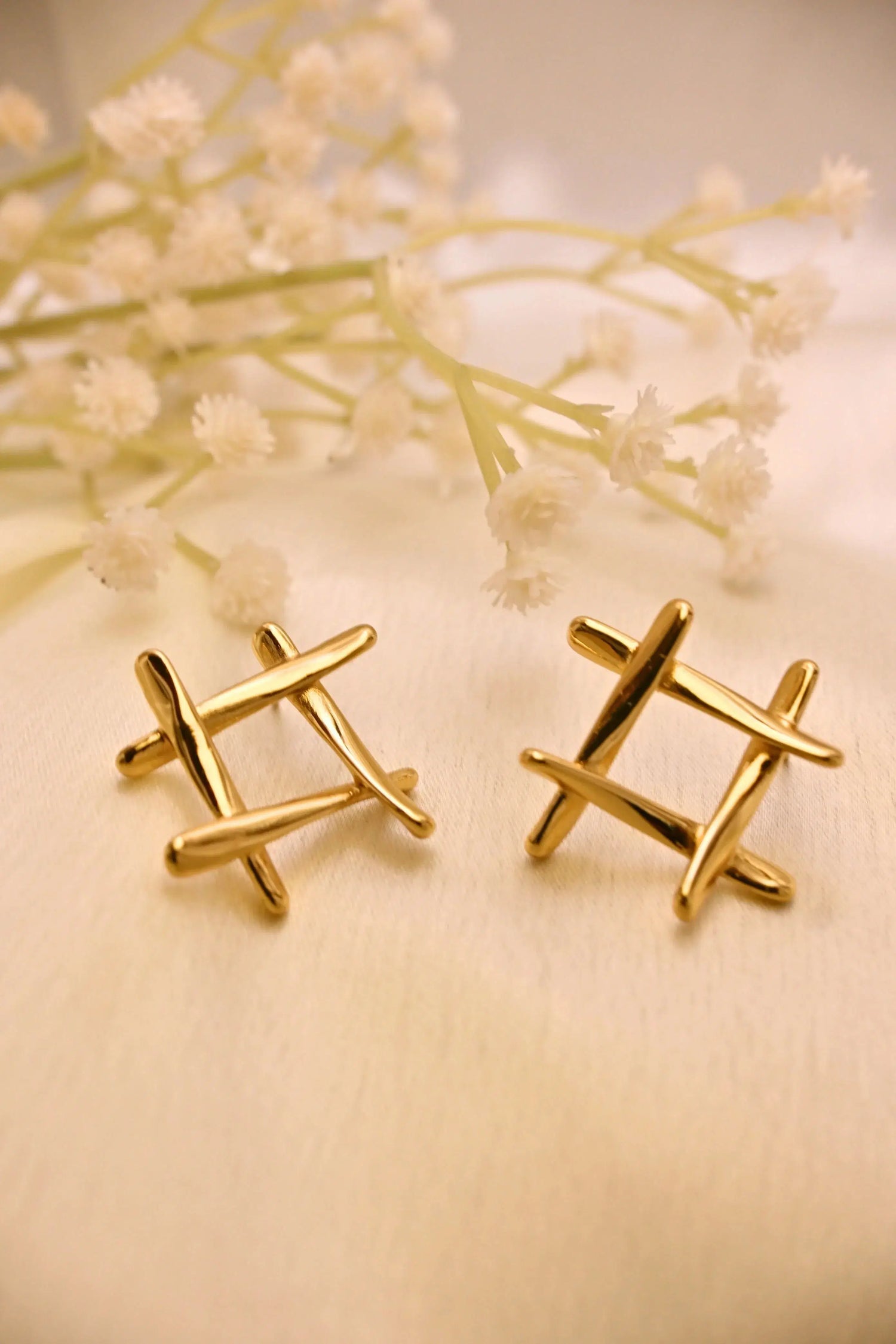 Pair of modern gold stud earrings with crisscross design on cream fabric, floral decor