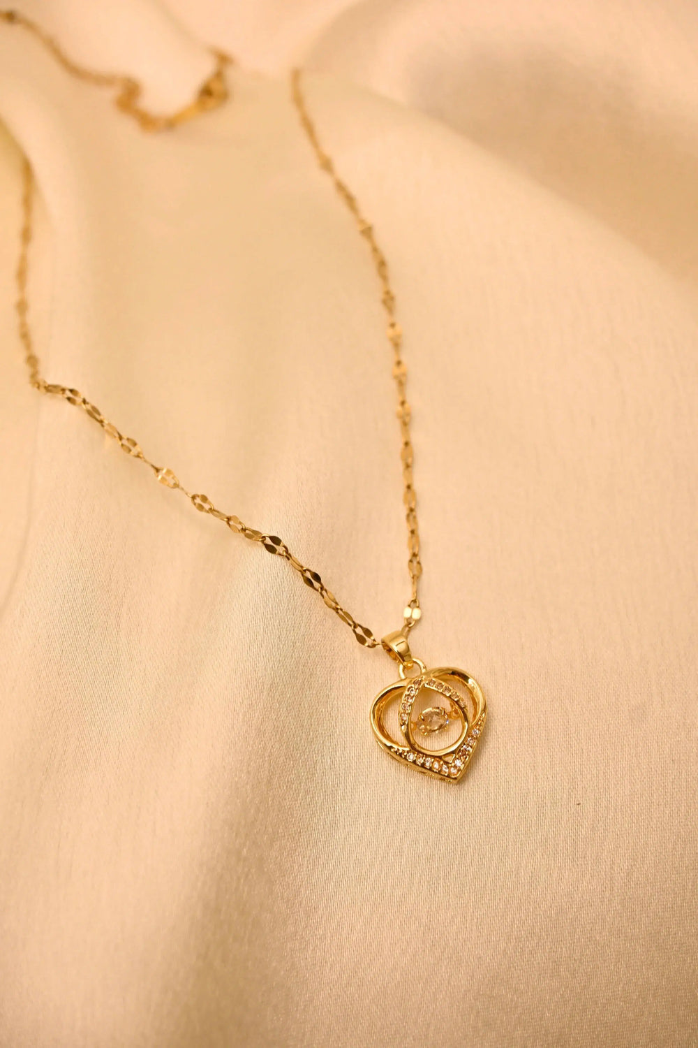 Gold heart pendant necklace with sparkling stones on a delicate chain, displayed on cream fabric