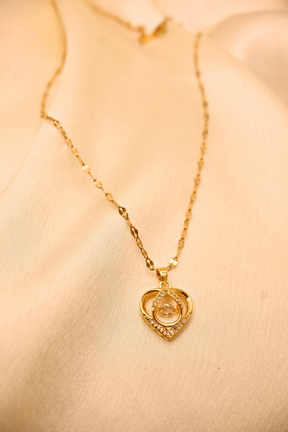 Gold chain necklace with a heart-shaped pendant featuring sparkling stones, displayed on a beige fabric.