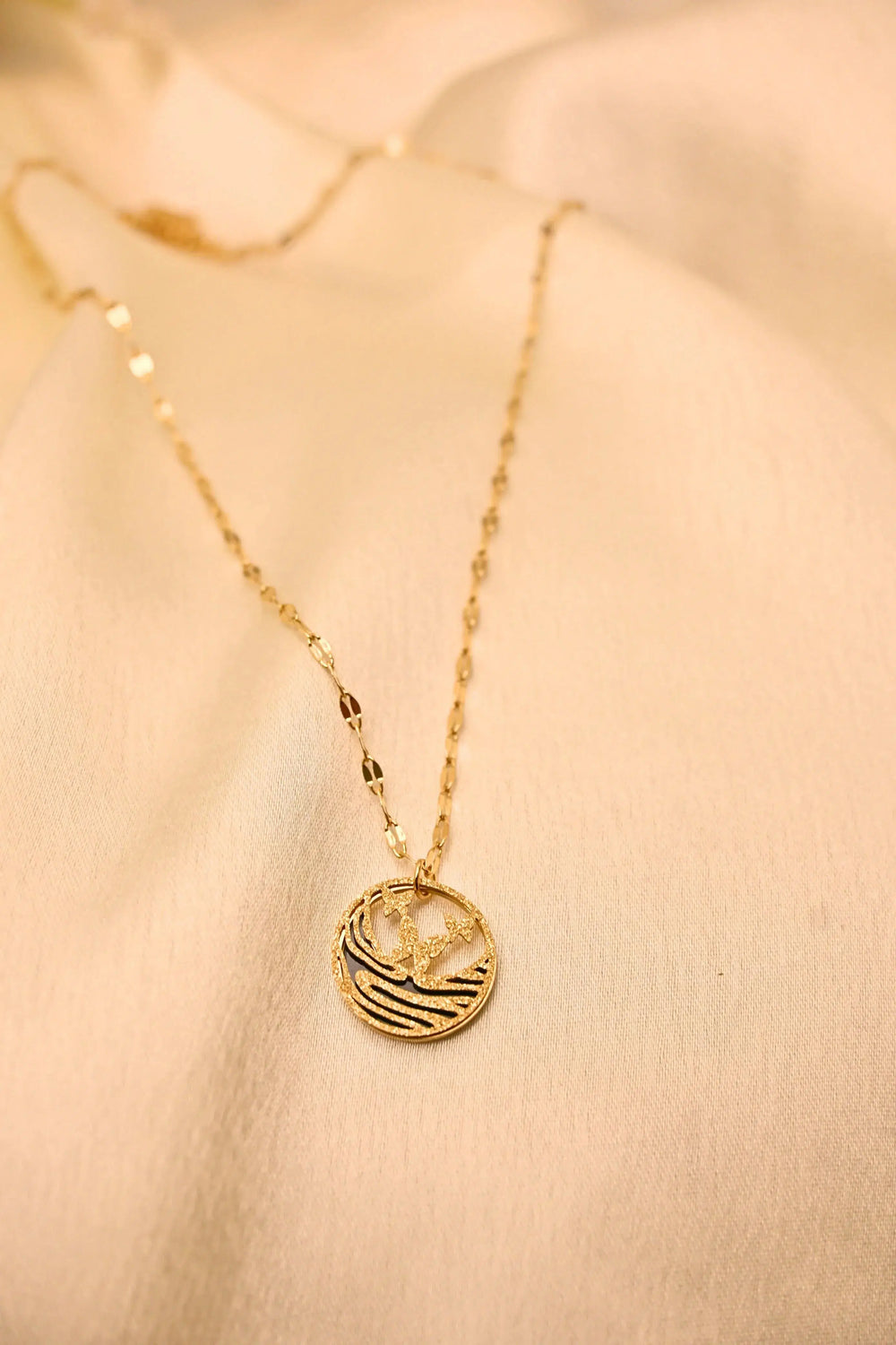 Gold chain necklace with a textured round pendant featuring a bird design on a cream fabric background