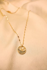 Gold chain necklace with a textured round pendant featuring a bird design on a cream fabric background