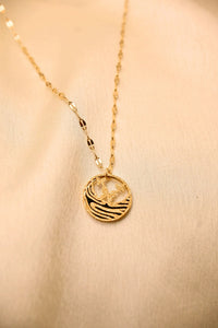 Gold pendant necklace with palm tree and wave design on textured chain, displayed on beige fabric