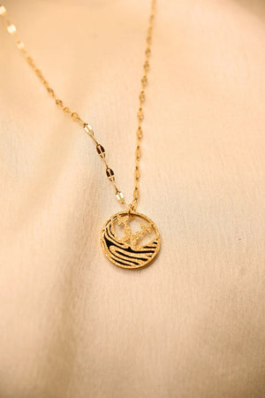 Gold pendant necklace with palm tree and wave design on textured chain, displayed on beige fabric