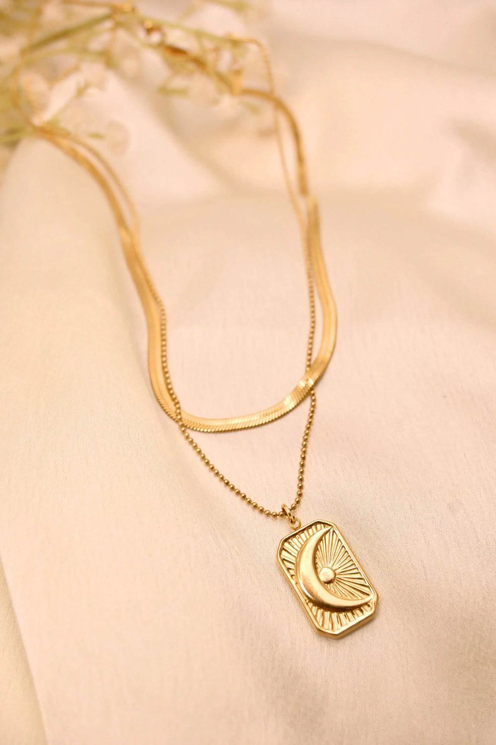 Layered gold necklace with rectangular moon pendant on beige fabric background