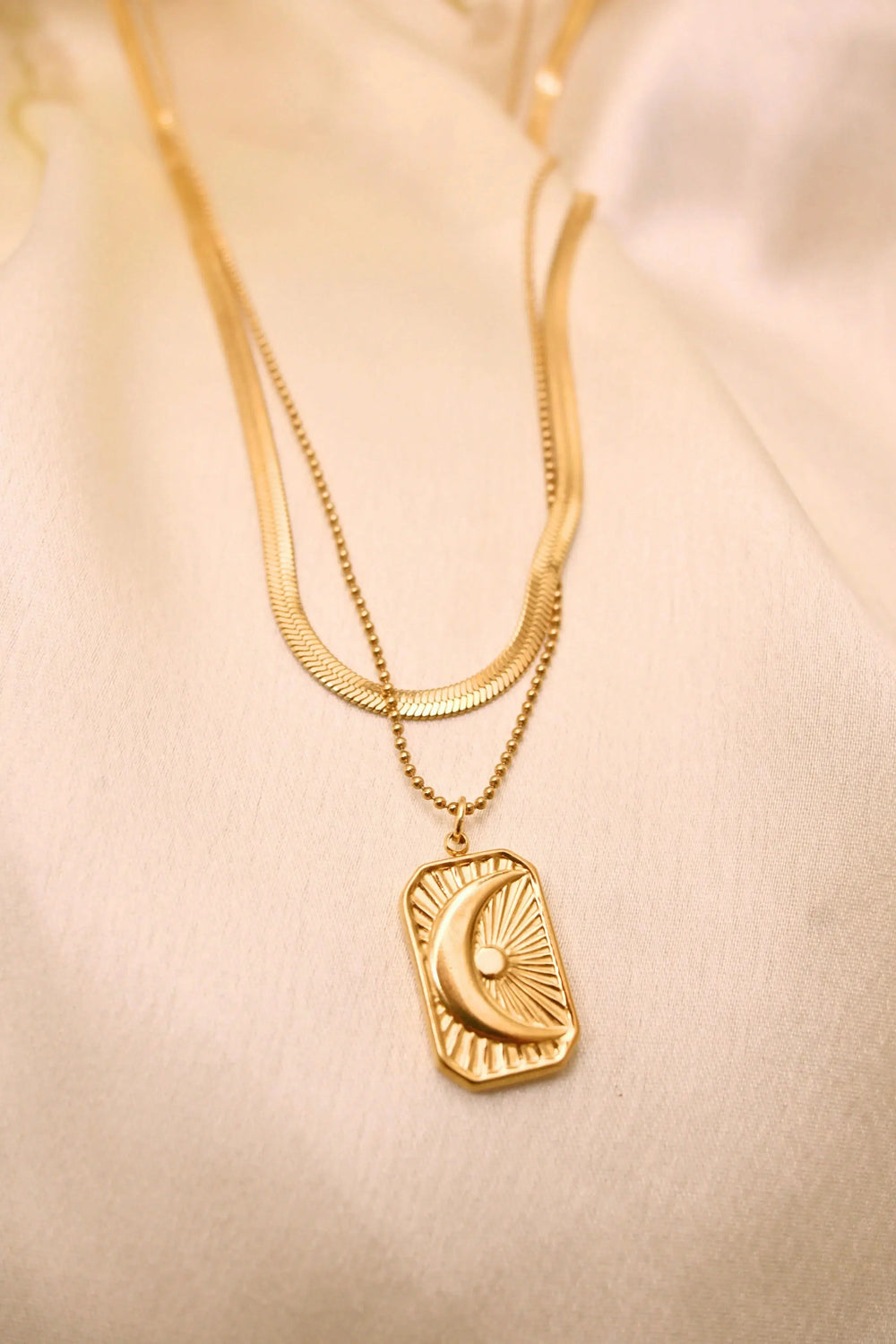 Layered gold necklace with crescent moon and sun pendant on cream fabric background