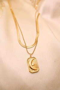 Layered gold necklace with crescent moon and sun pendant on cream fabric background