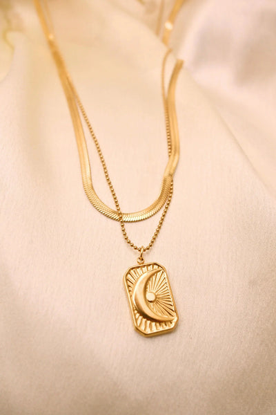 Layered gold necklace with crescent moon and sun pendant on cream fabric background
