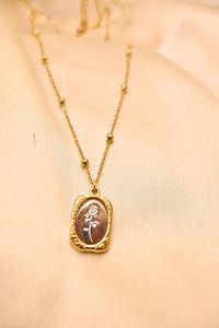 Gold pendant necklace with rose engraving on textured chain, displayed on cream fabric.