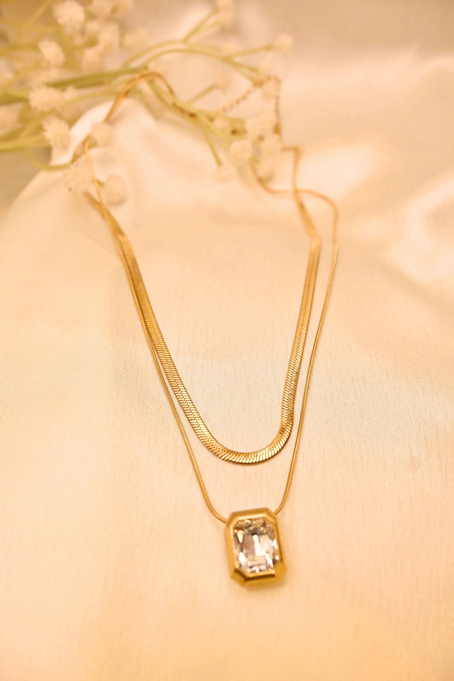 Layered gold necklace with a rectangular crystal pendant on soft beige fabric