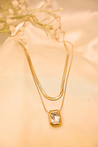 Layered gold necklace with a rectangular crystal pendant on soft beige fabric