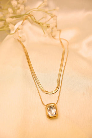 Layered gold necklace with a rectangular crystal pendant on soft beige fabric