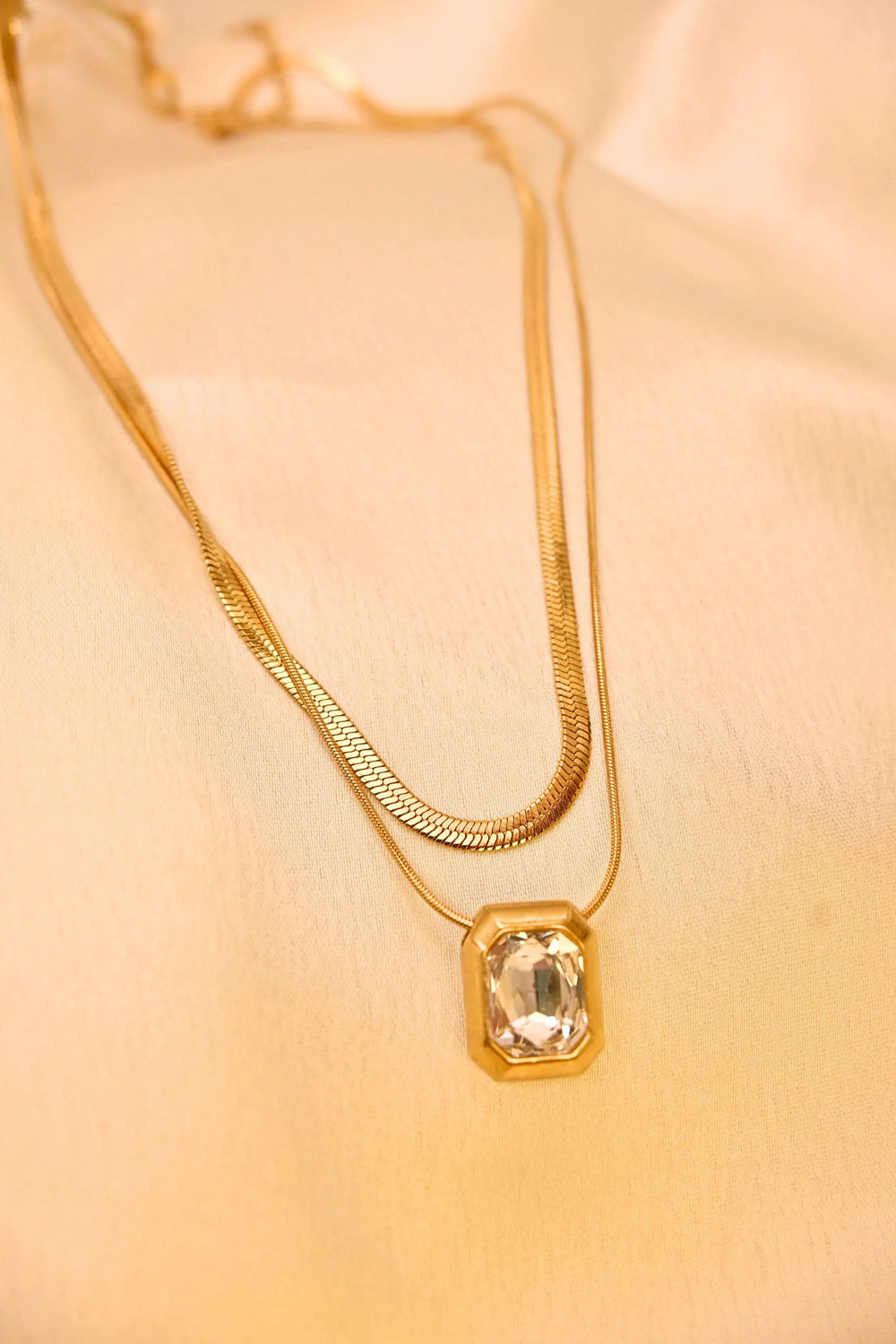 Gold layered necklace with clear rectangular gemstone pendant on cream fabric background